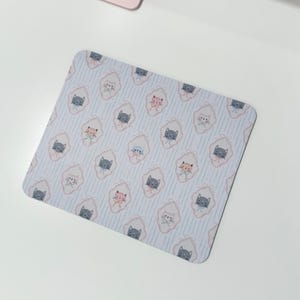 May include: A square mousepad with a repeating pattern of cartoon cats in pink, gray, and blue. The cats are inside a pink and white oval shape, set against a light blue striped background. The mousepad is on a white surface.