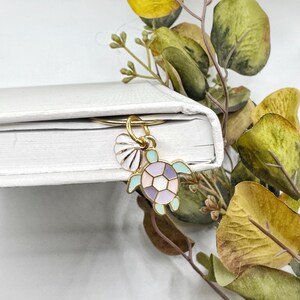 May include: A gold-colored metal bookmark with a sea turtle and seashell charm. The turtle has a pastel color scheme of light blue, lavender, and pink. The seashell is white with gold accents. The bookmark is attached to a white book.
