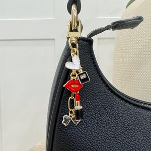 May include: A black handbag with a gold chain and charm. The charm features a red lip, a heart, a perfume bottle, and a lipstick.