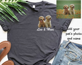 May include: A dark gray t-shirt with a white design featuring two golden retriever puppies and the text "Leo & Max". The t-shirt is shown on a white surface with a pair of brown sandals and blue jeans.