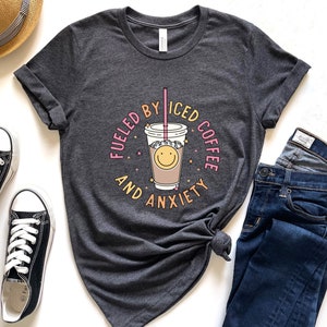 May include: A dark gray t-shirt with a graphic of a cup of iced coffee with a smiley face and the text "Fueled by Iced Coffee and Anxiety".