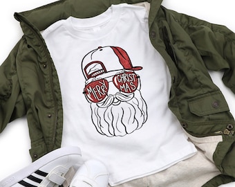 May include: A white t-shirt with a graphic of Santa Claus wearing sunglasses and a baseball cap, with the text "MERRY CHRISTMAS". The shirt is paired with an olive green jacket, white shorts, and white sneakers.