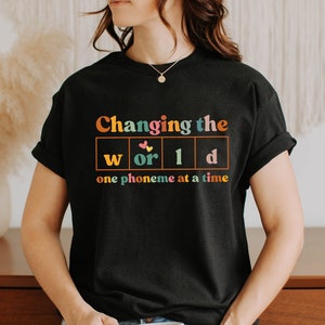May include: Black t-shirt with the text "Changing the world one phoneme at a time" in colorful retro font. The design includes a periodic table-style layout with the words "world" and "d" in boxes. A necklace is visible.