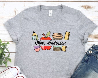 May include: Gray t-shirt with a graphic design of a red apple, pencil, coffee cup, and ruler. The text "Mrs. Anderson" is printed on a white banner below the graphic.