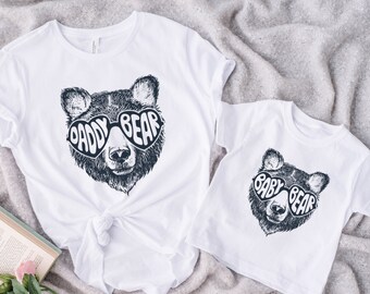 May include: Two white t-shirts with a bear wearing heart-shaped sunglasses. The larger shirt says "Daddy Bear" and the smaller shirt says "Baby Bear".