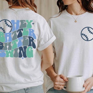 May include: Two white t-shirts with a baseball theme. One shirt has a blue baseball graphic on the front and the other has a blue baseball graphic and the text "Hey Batter Batter Swing" on the back.