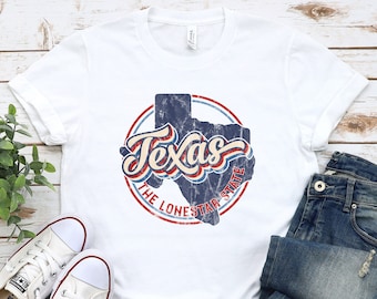 May include: White t-shirt with a distressed graphic of the Texas state outline, the word "Texas", and "The Lone Star State" in a circular design. The shirt is paired with blue jeans and white Converse sneakers.