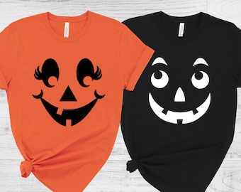 May include: Two Halloween-themed t-shirts. One is orange with a black jack-o'-lantern face featuring eyelashes, and the other is black with a white jack-o'-lantern face. Both shirts have rolled sleeves and are knotted at the bottom.