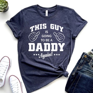 May include: A navy blue t-shirt with white text that reads "This guy is going to be a Daddy Again!" with thumbs up symbols on either side of the text.