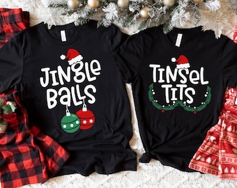 May include: Two black t-shirts with Christmas-themed text and graphics. One shirt says "JINGLE BALLS" with ornaments, the other says "TINSEL TITS" with a wreath. Red and black plaid pajama pants and red Christmas-themed pajama pants are also visible.