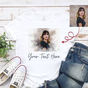 May include: White t-shirt with a watercolor image of a person and the text "Your Text Here". The t-shirt is laid out on a wooden surface with a pair of white sneakers, blue jeans, and a smaller image of the person in the top right corner.