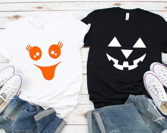 May include: Two t-shirts, one white and one black, with Halloween-themed designs. The white shirt features an orange ghost face, and the black shirt has a jack-o'-lantern face. Both are paired with blue jeans and white sneakers.