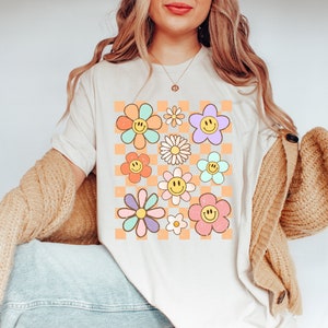 May include: A white t-shirt with a checkered pattern featuring colorful flowers with smiley faces. The flowers are in shades of pink, blue, yellow, and green.