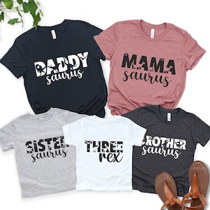 May include: Five t-shirts with dinosaur-themed designs. The shirts are navy, mauve, gray, white, and charcoal. Text includes "Daddy Saurus", "Mama Saurus", "Sister Saurus", "Three Rex", and "Brother Saurus".