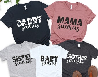 May include: A set of five t-shirts with a dinosaur theme. The shirts feature the words "Daddy saurus", "Mama saurus", "Sister saurus", "Baby saurus", and "Brother saurus".