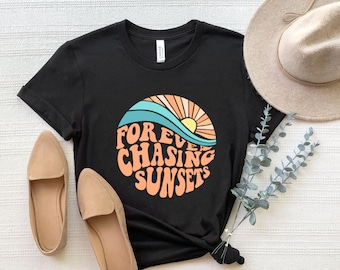 May include: Black t-shirt with a retro design featuring a sunset, waves, and the text "Forever Chasing Sunsets" in orange and teal. The shirt is paired with tan loafers and a beige hat.
