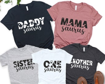 May include: Five t-shirts in various colors, each with a dinosaur-themed design. The shirts read "Daddy Saurus," "Mama Saurus," "Sister Saurus," "One Saurus," and "Brother Saurus" in white and black lettering. A pair of brown sandals is visible.