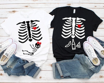 Puede incluir: Dos camisetas, una blanca y otra negra, cada una con un diseño de caja torácica de esqueleto. La camiseta blanca tiene un bebé esqueleto y un corazón rojo. La camiseta negra tiene un corazón rojo y gráficos de comida. Ambas camisetas se combinan con vaqueros y zapatillas blancas.