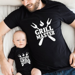 May include: A black t-shirt with the text "Grill Master" and a graphic of crossed grilling forks and flames. A matching black baby onesie with the text "Future Grill Master" and a similar graphic.