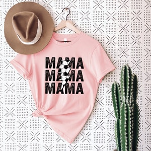 May include: Pink t-shirt with a black lightning bolt graphic and the text "MAMA" repeated three times. The lightning bolt is made up of black and white soccer balls.
