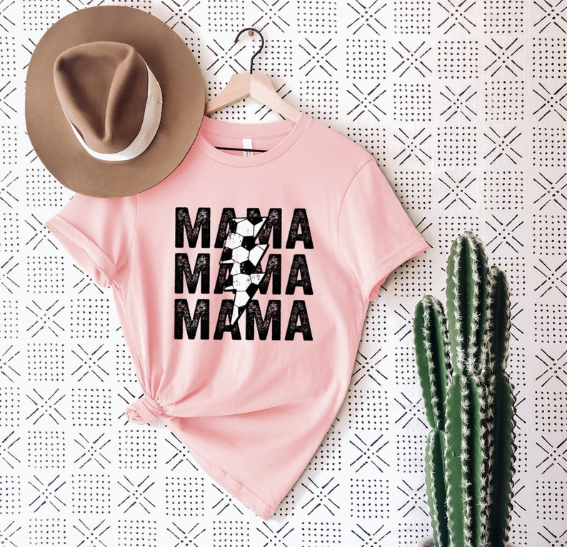 May include: Pink t-shirt with a black lightning bolt graphic and the text "MAMA" repeated three times. The lightning bolt is made up of black and white soccer balls.
