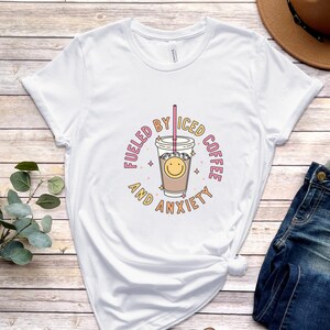 May include: White t-shirt with a graphic of a cup of iced coffee with a smiley face and the text "Fueled by Iced Coffee and Anxiety".