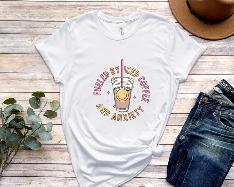 May include: White t-shirt with a graphic of a cup of iced coffee with a smiley face and the text "Fueled by Iced Coffee and Anxiety".