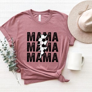 May include: A mauve t-shirt with a distressed black graphic of the word "MAMA" repeated three times with a lightning bolt made of soccer balls in the center.