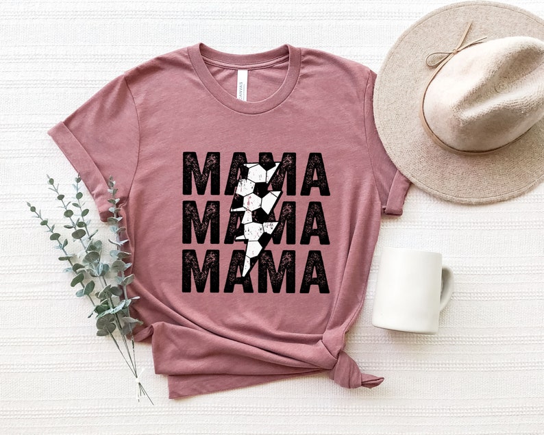 May include: A mauve t-shirt with a distressed black graphic of the word "MAMA" repeated three times with a lightning bolt made of soccer balls in the center.