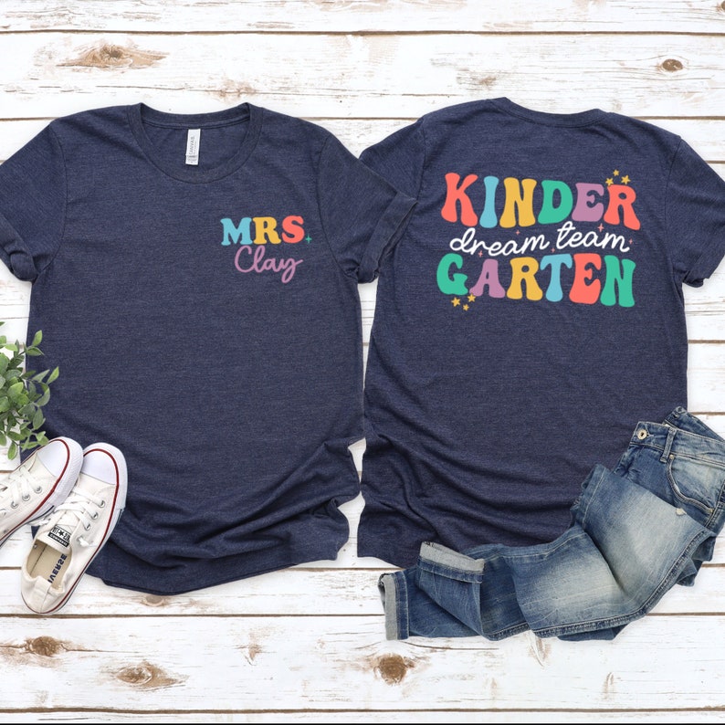 May include: Two navy blue t-shirts with text. The front shirt says "MRS Clay" in colorful letters. The back shirt says "Kinder dream team Garten" in colorful letters with stars.