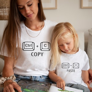 May include: White t-shirts with black keyboard key graphics. One shirt reads "Ctrl + C COPY" and the other reads "Ctrl + V PASTE". The shirts are worn by a mother and child, suggesting a matching set.