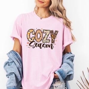 May include: A light pink t-shirt with the words "COZY Season" in a decorative font. The letters are filled with fall-themed patterns, including pumpkins, plaid, and zigzags. The shirt is part of a fall-themed clothing collection.