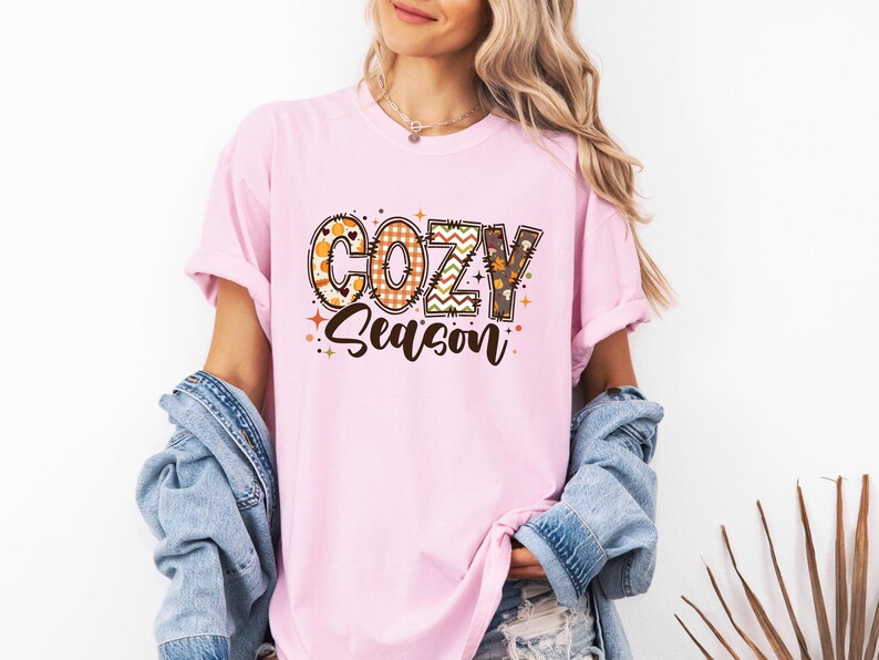 May include: A light pink t-shirt with the words "COZY Season" in a decorative font. The letters are filled with fall-themed patterns, including pumpkins, plaid, and zigzags. The shirt is part of a fall-themed clothing collection.