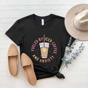 May include: Black t-shirt with a graphic of a cup of iced coffee with a smiley face and the text "Fueled by Iced Coffee and Anxiety".