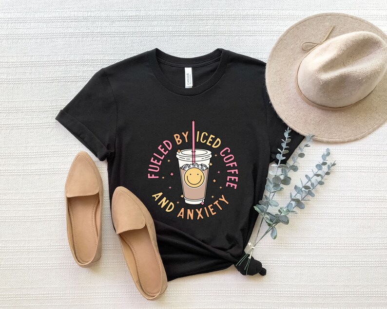 May include: Black t-shirt with a graphic of a cup of iced coffee with a smiley face and the text "Fueled by Iced Coffee and Anxiety".