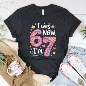 May include: Dark grey t-shirt with the text "I was now 67 I'm" in a colourful design. The number 67 is pink with hearts and stars. The shirt is paired with jeans, trainers, and a mug.