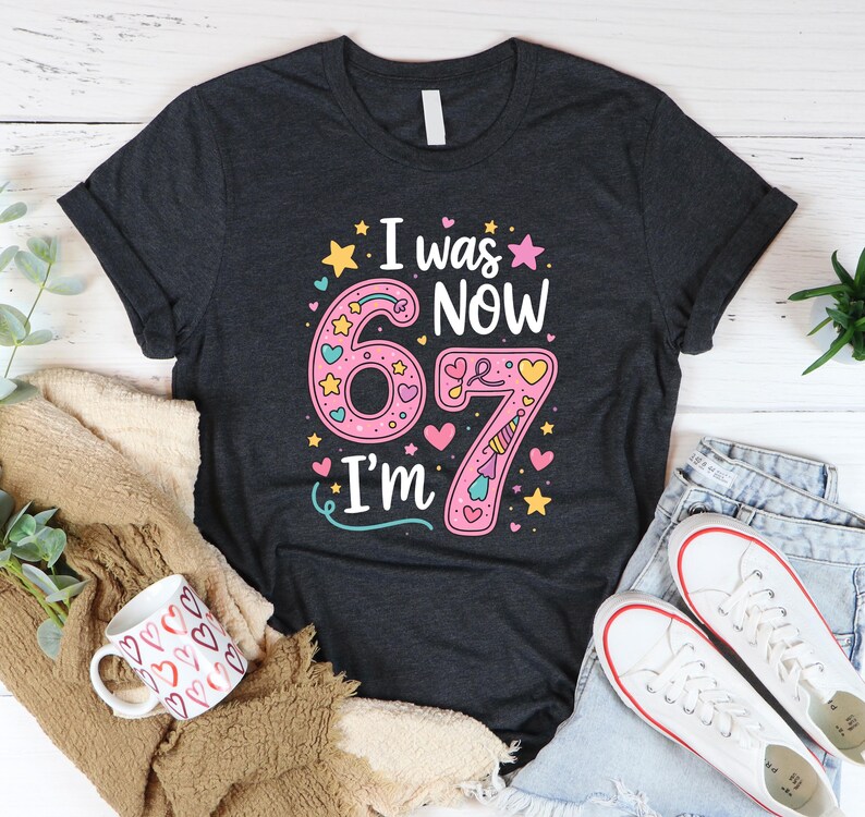 May include: Dark grey t-shirt with the text "I was now 67 I'm" in a colourful design. The number 67 is pink with hearts and stars. The shirt is paired with jeans, trainers, and a mug.