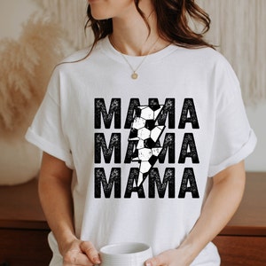 May include: White t-shirt with a black and white graphic of a soccer ball and lightning bolt. The text "MAMA" is repeated three times in a distressed font.
