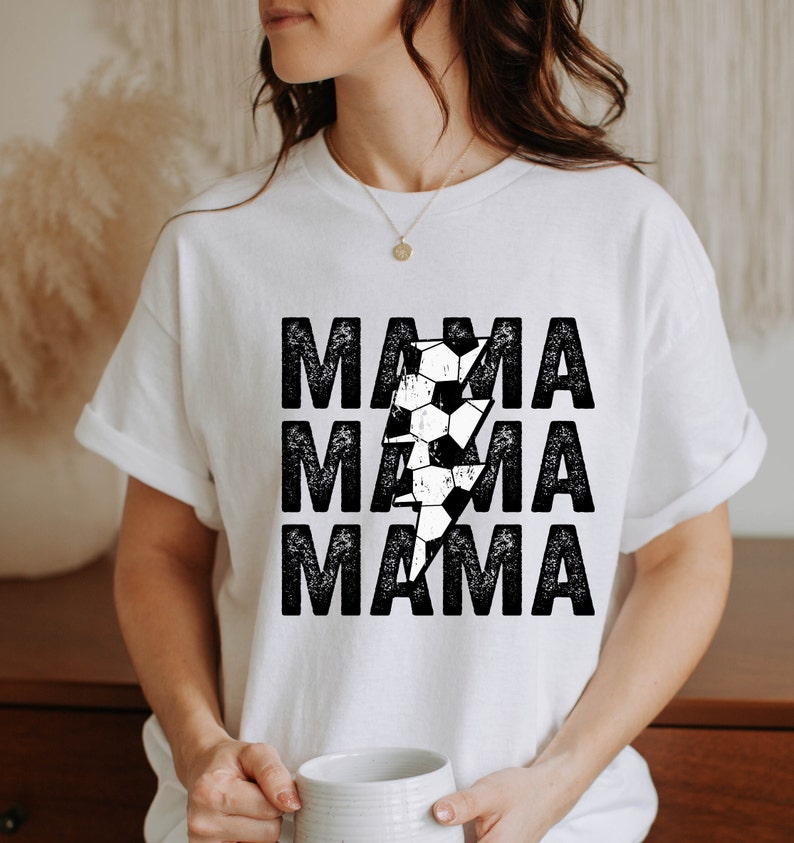 May include: White t-shirt with a black and white graphic of a soccer ball and lightning bolt. The text "MAMA" is repeated three times in a distressed font.