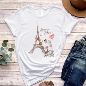 May include: White t-shirt with a graphic of the Eiffel Tower, flowers, and two people riding a bicycle. The text "Bonjour Paris!" is printed on the shirt.