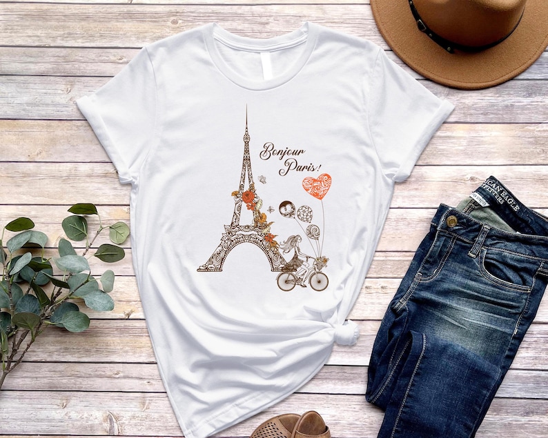 May include: White t-shirt with a graphic of the Eiffel Tower, flowers, and two people riding a bicycle. The text "Bonjour Paris!" is printed on the shirt.