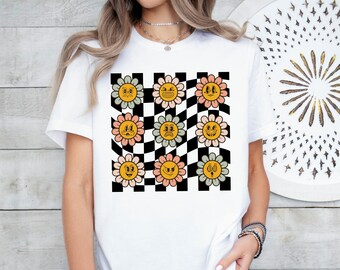 May include: White t-shirt featuring a retro-style design of smiling flower faces on a black and white checkered background. The flowers have yellow, pink, and green petals with various facial expressions. The shirt is a casual, everyday wear item.