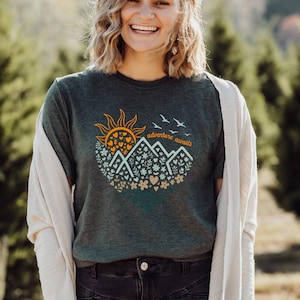 May include: Dark green t-shirt featuring a circular graphic with a sun, mountains, flowers, and the text "adventure awaits." The sun is orange with heart-shaped details. The mountains are white, and the flowers are in shades of green and beige.