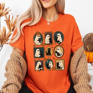 Puede incluir: Camiseta naranja con un dise&ntilde;o tem&aacute;tico de Halloween. El dise&ntilde;o incluye ilustraciones enmarcadas de fantasmas y gatos negros. Los marcos tienen varias formas y est&aacute;n decorados con hojas y otros elementos de temporada. La camiseta es de material suave.