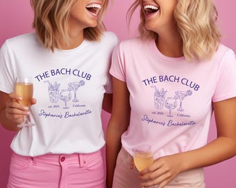 May include: Two women wearing white and pink t-shirts with the text "THE BACH CLUB" and "Stephanie's Bachelorette" printed on them. The shirts also feature illustrations of cocktails. Both women are holding glasses of champagne.