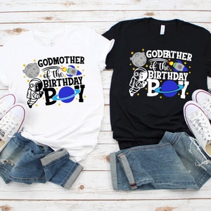 May include: Two t-shirts, one white and one black, with a space-themed design. The white shirt says "Godmother of the Birthday Boy" and the black shirt says "Godfather of the Birthday Boy". Both shirts feature an astronaut, a planet, a rocket, and stars.