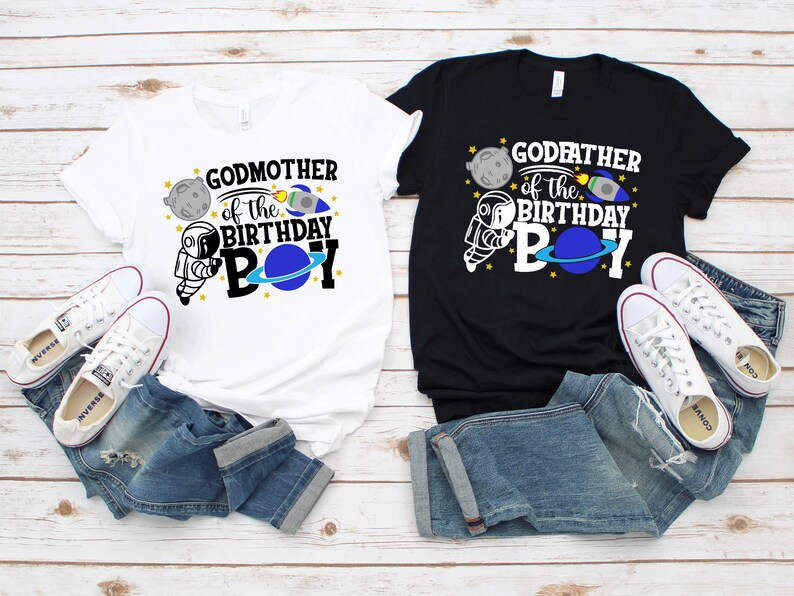 May include: Two t-shirts, one white and one black, with a space-themed design. The white shirt says "Godmother of the Birthday Boy" and the black shirt says "Godfather of the Birthday Boy". Both shirts feature an astronaut, a planet, a rocket, and stars.