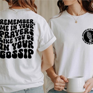 May include: Two white t-shirts with black text. The front of one shirt has a graphic of a praying hand in a circle with the text "Remember me in your prayers like you do in your gossip". The back of the other shirt has the text "Remember me in your prayers like you do in your gossip" in a retro font.