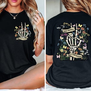 May include: Black t-shirt featuring a graphic of a skeleton hand making a peace sign surrounded by flowers. The text "Good Energy is Contagious" is on a banner below the hand.