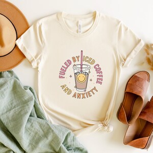 May include: A white t-shirt with a graphic design of a cup of iced coffee with a smiley face and the text "Fueled by Iced Coffee and Anxiety".
